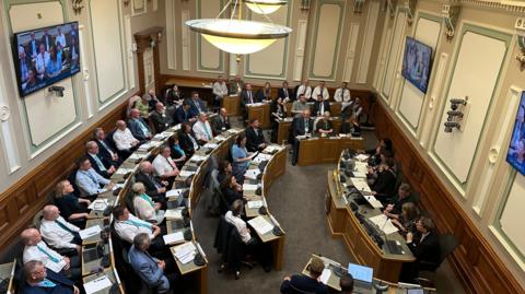 Looking down into a council chambers, circular rows of desks with people sat in smart clothes. In front of them a smaller desk with five people sat facing them. Screens are on the walls of the room and there are overhanging lights.