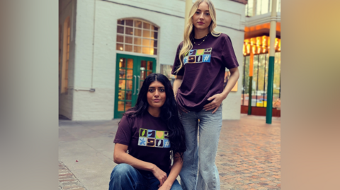 Two girls model the purple T-shirts in a courtyard with a white painted building behind them. One girl with long, dark hair has one knee on the ground while the other girl, who has long, blonde hair is stood to her side.