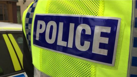 The back view of a British police officer's vest. It s bright yellow with a blue rectangle and white writing on it that says POLICE