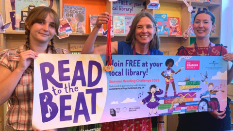 Lexie Le Poidevin, Kate Knivett, Reinet Smith are standing in the children's library with books behindt hem. They are holding a banner saying Read to the Beat, join free at your local library. Kate who is in the middle is holding up medals on red ribbons. 