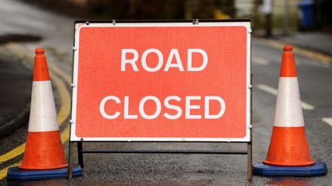 A close up of a red sign with the words "road closed" printed on it in a white font. Two orange cones sit either side of the sign. The sign is speckled with rain water.