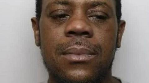 Anthony Blackwood with stubble and dark hair. He is looking away from the camera to the right and has a serious expression. It is his mugshot so he is stood in front of a grey background and is wearing what appears to be a grey top.