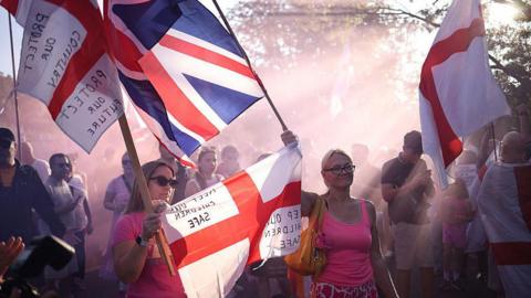 A crowd of protesters holding English flags and Union Jack flags at a protest in Epping. Two women in pink tops are in the foreground, while the crowd stands slightly shrouded by red mist from a flare.