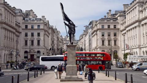 The statue is seen in a wide view of the street with crowds and a bus