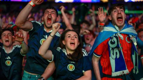 Fans celebrate in the OVO Hydro as Scotland score against Haiti. In the foreground are three men and one woman, all cheering and wearing Scotland strips. Rows of supporters can be seen behind them.