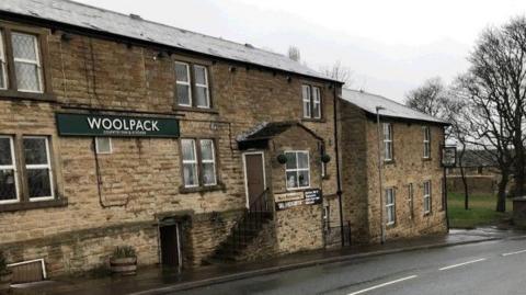 An old, large brick pub with a green sign on the front reading 'Woolpack'. There is a small set of stairs leading to the front door. 