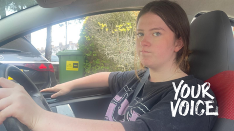 Beatrix sat in the driver's seat of her car, looking at the passenger who is taking a photo of her with a slightly grumpy expression on her face. She has shoulder-length black hair and is wearing a black T-shirt with a purple pattern on the front.