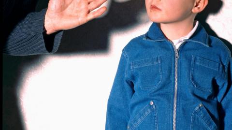 A person in a grey suit lifts their hand in front of a young child's face. The child's face is not shown. The child is wearing a denim zipped jacket and a white shirt. They are standing in front of a white wall.