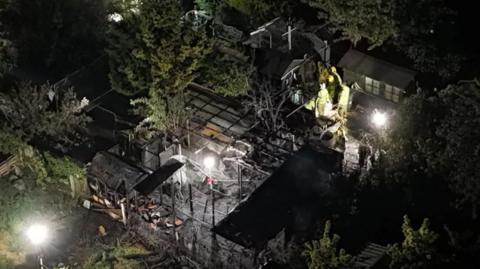 Charred remains for gardens seen at night with several firefighters among burnt out outbuildings.