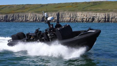 A black boat with two police officers travels quickly through the water near some cliff faces.