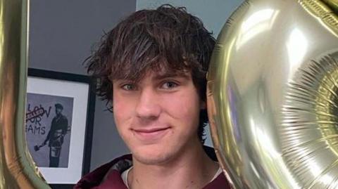 A man with dark hair is looking at the camera. A grey background is behind him on the left.
