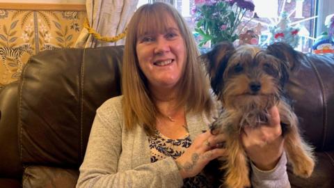 A woman sat on a sofa smiling holding a Yorkshire Terrier puppy in her hands that is brown and has black ears