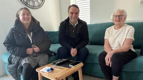 A picture of three people sat on a green sofa. There is a wooden table in front. One woman has white hair and black glasses. She is wearing a white top and black trousers. A man is sat in the middle wearing dark coloured clothes and a woman is sat next to him with brown hair and black coat. All are smiling.