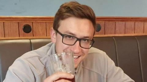 Kamil Grygieniec smiling at the camera with a cream top. He is holding a glass of beer and has glasses and light brown hair.