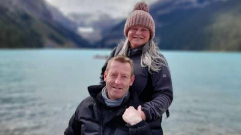 A man is seated in a wheelchair, wearing a black jacket. Behind him is a woman resting a hand on his shoulder. She is dressed in a black jacket and has long blonde hair and is wearing a knitted hat. Both are smiling. Behind them is a large expanse of water surrounded by mountains.