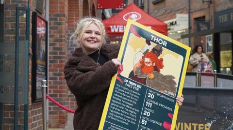 A woman with blonde hair wearing a brown fluffy coat holds a giant yellow Top Trumps card featuring the Norse god Thor.