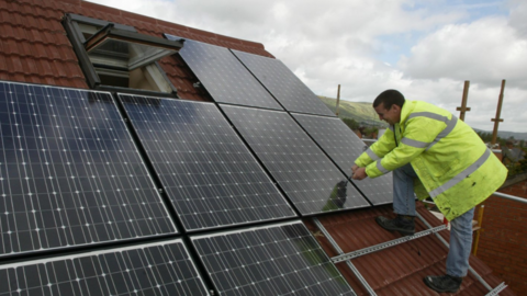 The picture shows a person wearing a bright yellow high-visibility jacket working on the installation of solar panels on a sloped, red-tiled roof. Several large black photovoltaic panels are already mounted in neat rows, and the person appears to be securing one of the panels using tools. There is an open roof window near the top left of the image, and metal mounting rails are visible beneath the panels. In the background, scaffolding is set up along the roof edge, and the sky is partly cloudy with green hills visible in the distance.