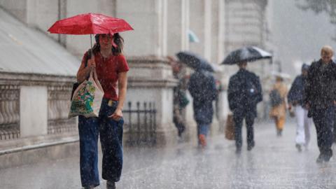 People on a rainy London street. In foreground person wearing red top with red open umbrella and bag.  