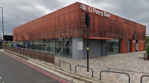 The corner of a train station building, which is part brown and part grey, with large glass windows and doors at the bottom. At the top it has a railway sign next to the words Perry Barr in white.