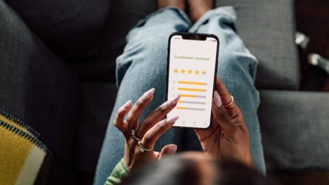 Stock photo shows an overhead view of a person holding a smartphone with a screen saying 'customer feedback' and a star rating interface, as they sit on the sofa at home.