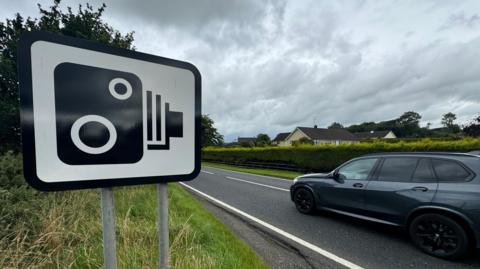 Image of a speed camera warning sign with a car driving past it. 