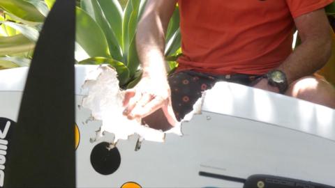 A shark teeth-sized bite in a wind surfboard next to a man with an orange shirt