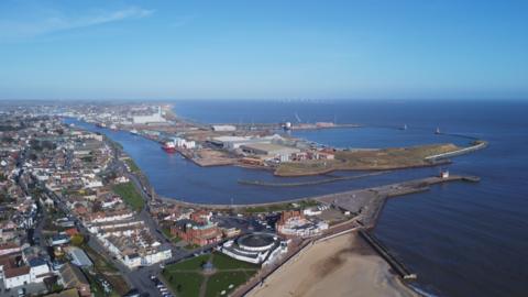 A drone image shows the mouth of a river and a large port area, with housing surrounding it.