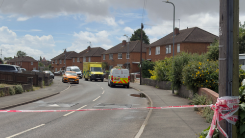 A residential street with police tape across the front. In the distance you can see several vehicles including an ambulance and a police van.