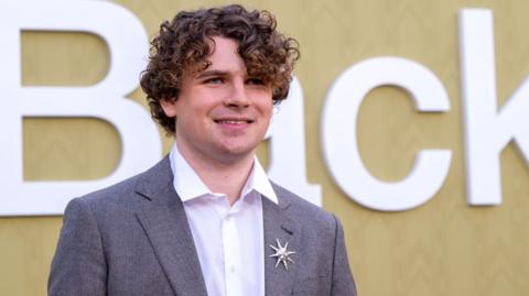 A poised, sharply dressed figure stands against a warm tan backdrop marked by large white lettering. The person has thick, curly hair that frames their face. They’re wearing a light grey suit jacket over a crisp white shirt, the collar open for a modern, unfussy finish. A small star‑shaped lapel pin catches the light on the left side of the jacket. The backdrop, with its oversized white characters (“B” and “ck” visible), suggests a formal event or premiere, the kind where attendees are photographed on arrival.