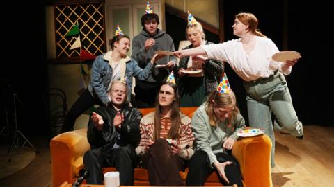 Three people are sat on a sofa, with four standing behind them. they are all wearing party hats and holding paper plates. 