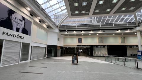 The picture shows the empty Nicholsons shopping centre with the shutters for businesses pulled down. A lone 'A' board stands in the middle of the shot with the centre's grey stone paved walkway devoid of shoppers.  