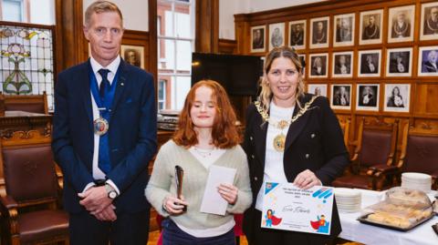 Evie, a teenager with ginger hair, with her eyes closed holding the award. She is standing between Douglas Mayor Natalie Byron-Teare, who is wearing her official chain, and consort Andy Teare in the council chamber.