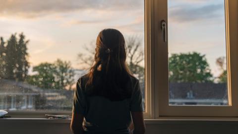 A young girl looks out of her bedroom window as the sun is setting. The view through the window is blurry but shows a residential area.