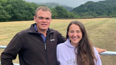 Sara Jenkins a Ioan Jones yn sefyll o flåen cae gyda coed a mynyddoedd yn ymestyn i'r gorwel