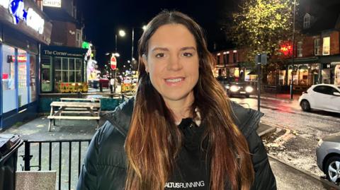 Sophie stands in a busy Hull street at night with lit-up bars and restaurants in the background. She is wearing a black hoodie with an Altus Running logo on the front and has long brown hair.
