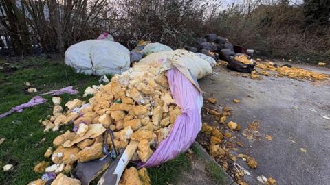 Several large bags of waste dumped on a supermarket car park. One of the bags coloured purple has split and what is believed to be foam loft installation has spilled out of it among other waste. The bags of rubbish are in front of trees.