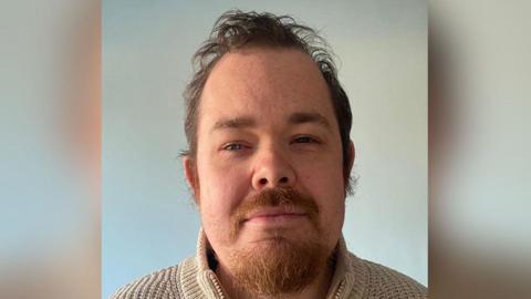 A head and shoulders shot of Lee Mercer smiling into the camera, against a plain background. He has short brown hair, a beard and moustache. He is wearing a beige sweatshirt with a zip.