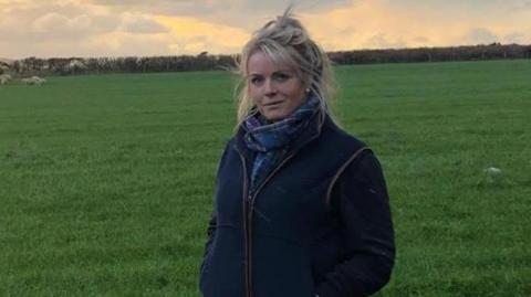 Kirree Kermode has blonde hair loosely tied up. She is wearing a blue Manx tartan scarf and a dark blue gilet over a dark coloured jumper. She is standing in a field at dusk.