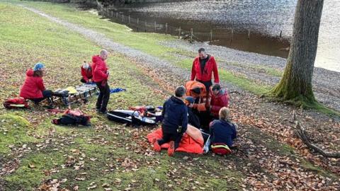 Several mountain rescue volunteers - dressed in red clothing - attend to a casualty on the path by Otterbield Bay, Derwentwater. There are lots of leaves scattered on the small slope and a stretcher has been prepared for the casualty. 