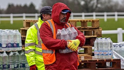 A man holding a pallet of water. There is also another man standing behind him. They are pallets of water stacked behind them.