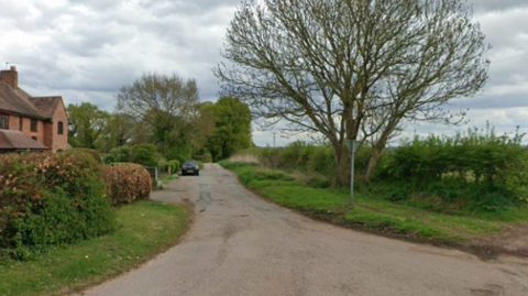 A country lane can be seen with a house on one side and a car parked in the lane with a tree and hedge on the opposite side.