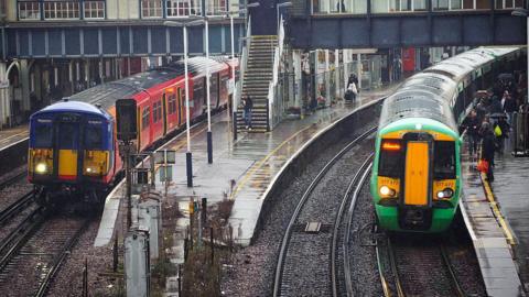 A blue and red South Western Railway train and a green Southern passenger train arrive at Clapham Junction station.