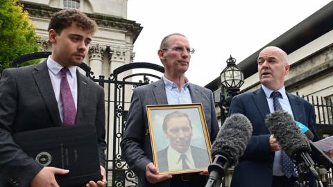Three men outside court. They wear suits. The man in the centre is holding a gold framed photograph of another man. There are microphones in front of them.