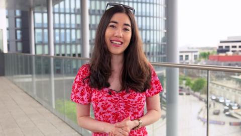 BBC Weather's Sabrina Lee, a young woman with dark hair, in a summer dress with sunglasses on her head smiles at the camera on a city rooftop.