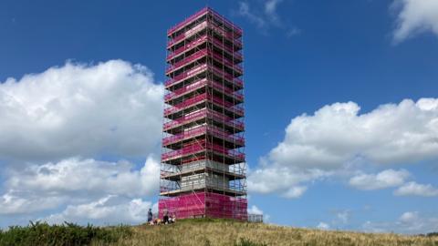 A tower in a field is covered in pink scaffolding. There is a blue sky with clouds in the background.