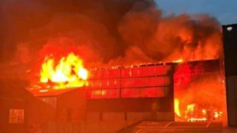 A large warehouse and side building are engulfed in flames. There is black smoke billowing from the fire. The picture is illuminated orange because of the fire.