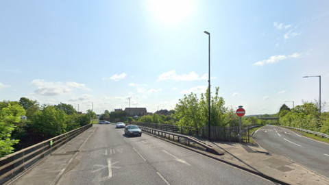 A two-lane road over the M1. It is a sunny day with blue skies. 