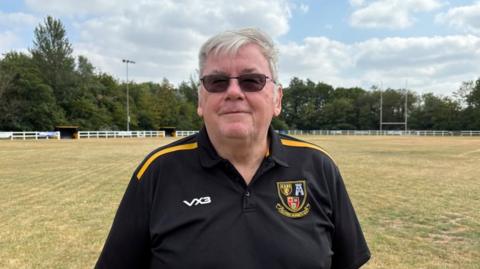 There is a man in a black polo shirt with yellow lines on the shoulders. He has two buttons undone. To the left of his shirt, it says the manufacturer VX3 and the right, the club's crest. He is wearing dark sunglasses and has short grey hair. Behind him, there is a pitch covered in brown grass. There is a rugby goal to his right and a floodlight to his left. 