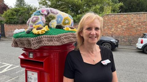 Mairi Wingate, the postmistress at Barton-upon-Humber. She is wearing a black top with a red and white name badge. She is standing next to a traditional red postbox with a knitted wool topper. A road with cars is visible in the background.