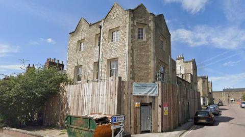 The Cricketers Pub building on the corner of a street with fencing around it and construction notices. There is a banner across the fencing entrance and a skip outside.